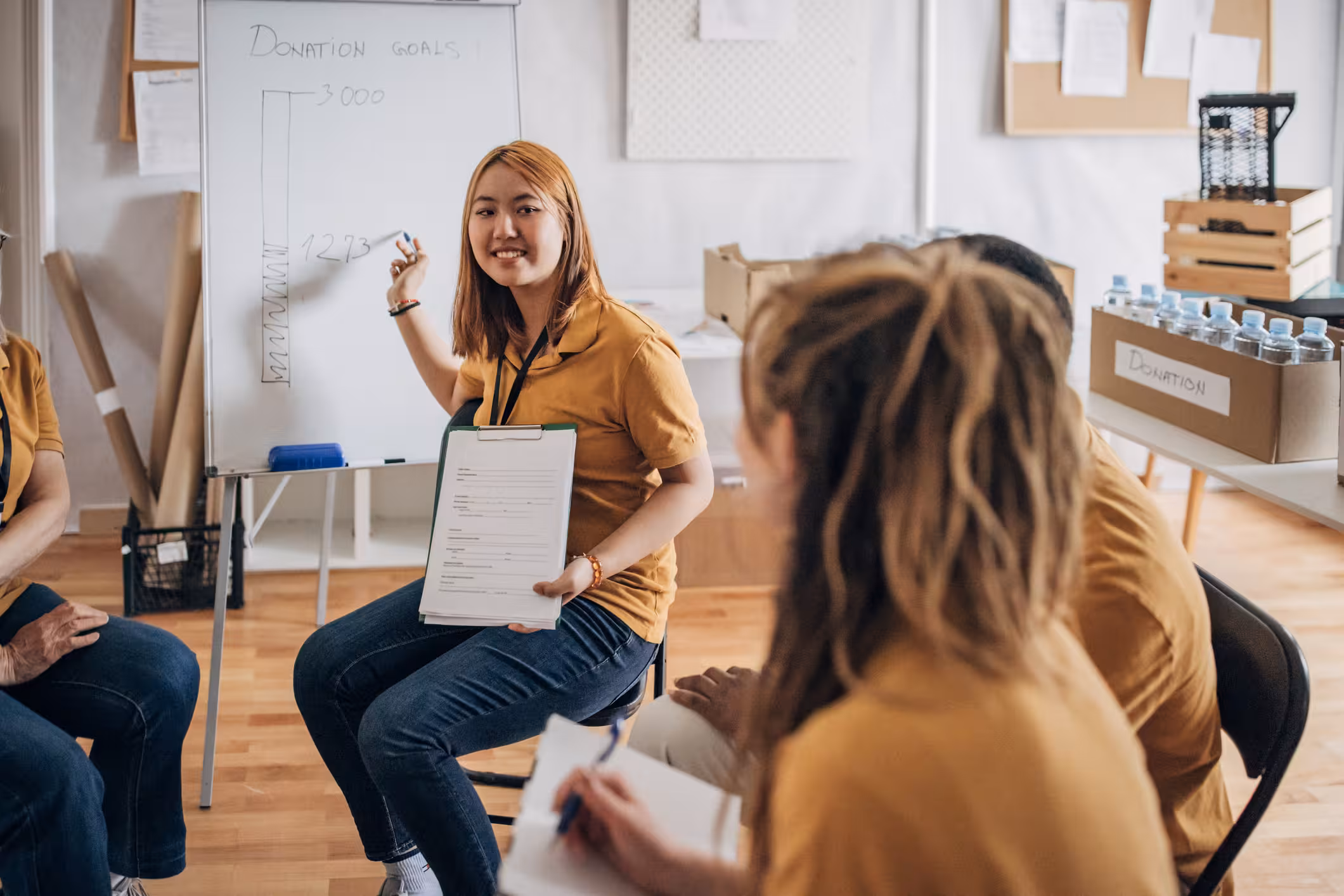 A woman in a yellow shirt presents donation goals on a whiteboard to a small group, holding papers while others listen and take notes in a bright room with boxes and water bottles.