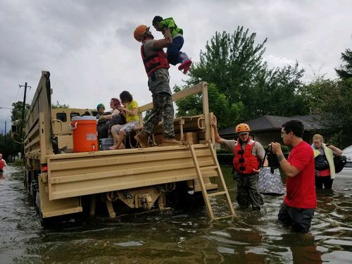 Several people in life jackets and helmets help families and children onto a military truck during a flood, with water covering the street and homes in the background. One rescuer lifts a child onto the truck.