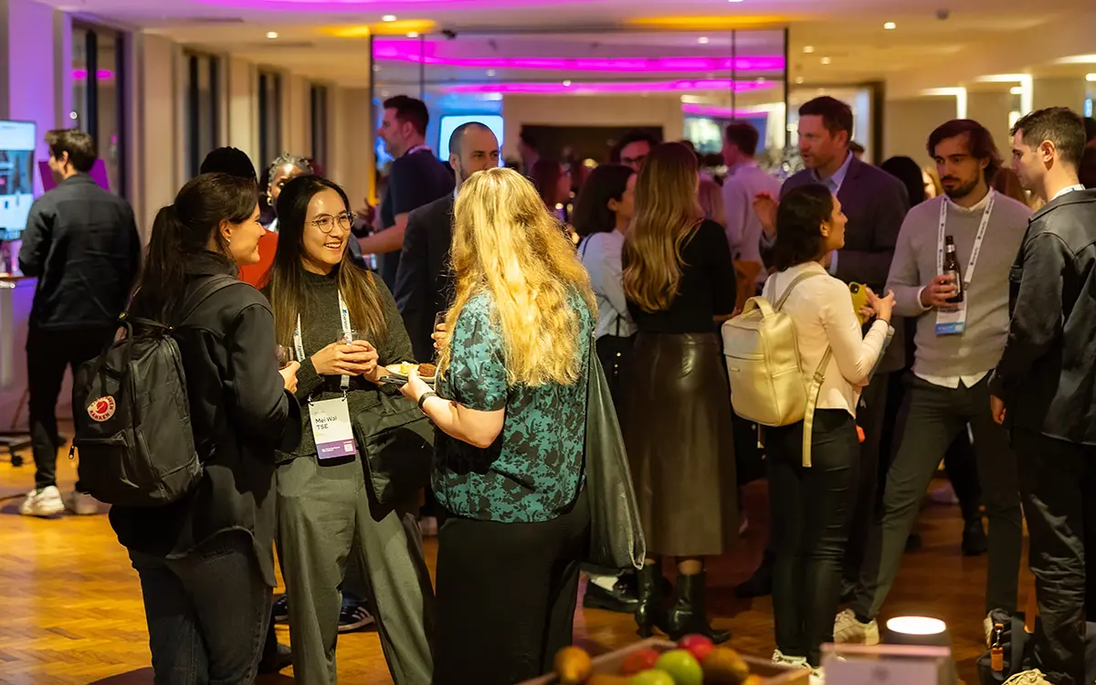 A group of people socialize and network in a brightly lit indoor event space, some holding drinks and wearing name badges, with tables, fruit bowls, and modern decor in the background.