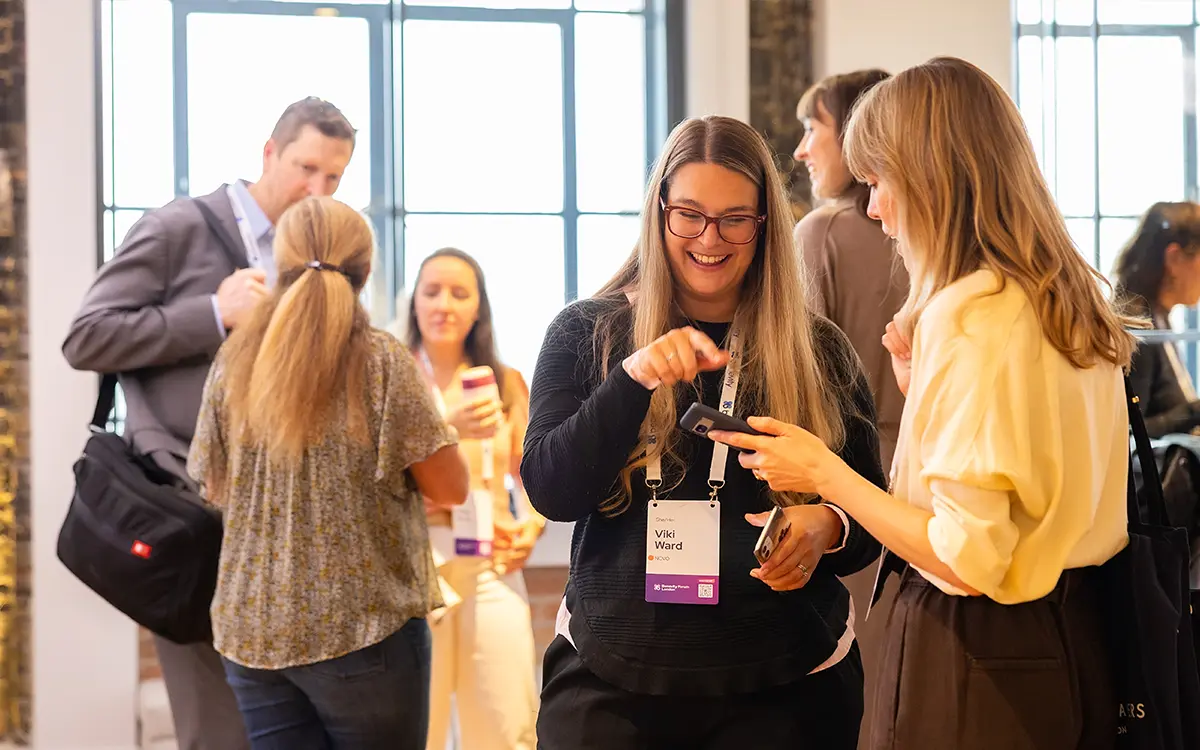 A group of people socialize indoors at an event. Two women in the foreground smile and look at a smartphone, while others in the background chat and hold drinks. Name badges are visible on some attendees.