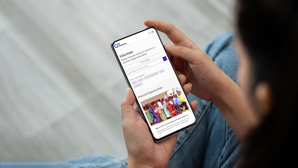 A person holds a smartphone displaying a volunteer search webpage with options to choose location, category, and keyword, along with featured volunteer opportunities and a group photo.