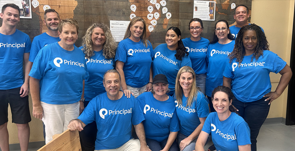 A group of thirteen adults, all wearing matching blue Principal t-shirts, smile and pose together.