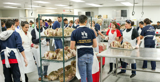 A group of people wearing aprons and gloves work together in a large kitchen, preparing and organizing multiple raw turkeys on metal racks and tables.