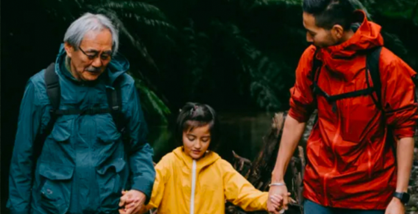 An older man, a young girl, and a man walk outdoors holding hands. They wear colorful jackets—blue, yellow, and red—amid a lush, green, forested background.