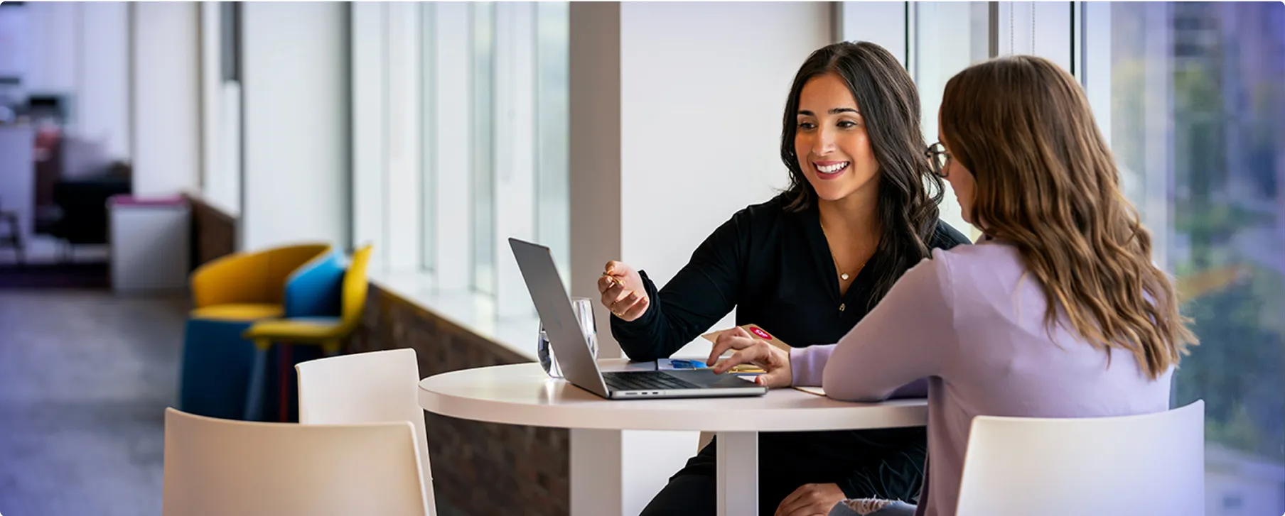 Two women sit at a round table by large windows, smiling and discussing work. One points at a laptop screen while the other listens attentively and types on the keyboard.