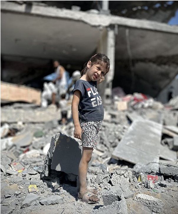 A young girl stands amid rubble and debris from destroyed buildings, wearing a black t-shirt and shorts. The background shows damaged structures and people searching through the ruins. The scene conveys devastation and loss.
