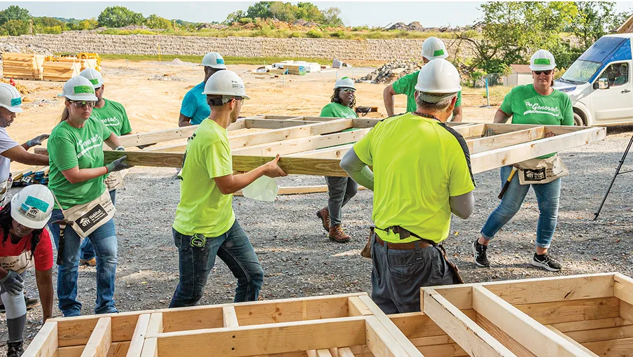 A group of people wearing hard hats and green shirts work together to carry wooden beams at a construction site, with partially built structures and construction materials visible in the background.