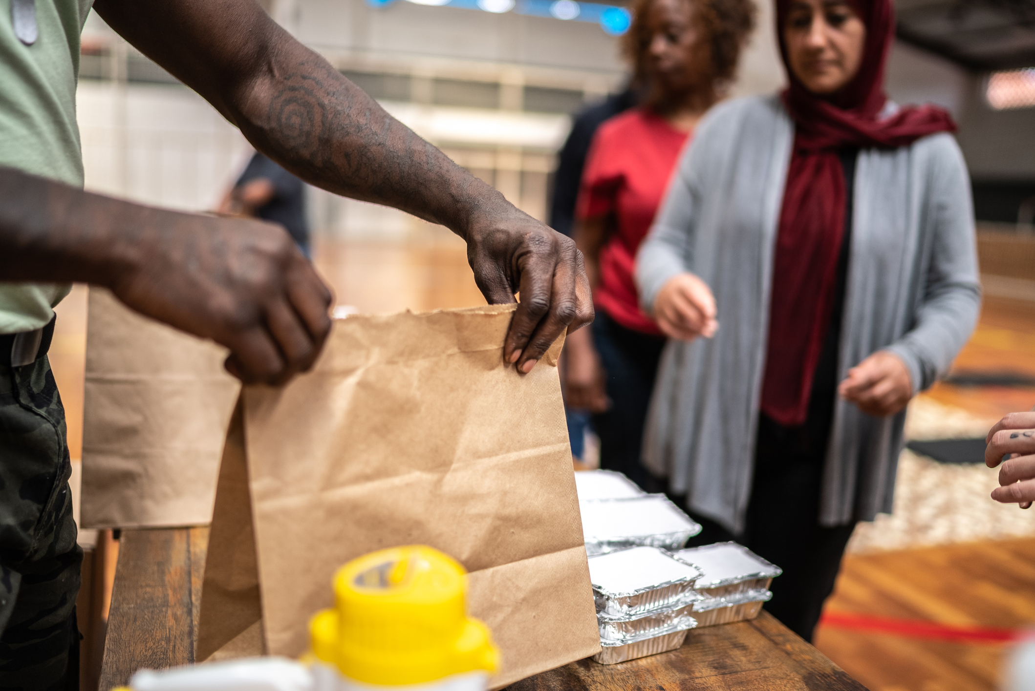 Several people stand around a table with food containers and brown paper bags, preparing or distributing meals in what appears to be a community center or gymnasium.