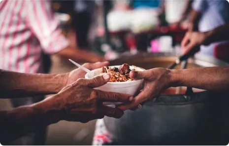 A person hands a bowl of food to another person, symbolizing food sharing or charitable meal distribution, with blurred people and food containers in the background.