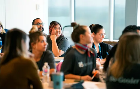 A group of people sit at tables in a bright room, listening attentively and taking notes during what appears to be a meeting or workshop. The atmosphere is focused and engaged.