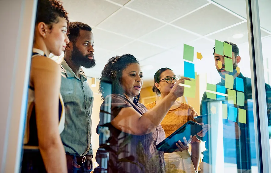 A group of five people stands by a glass wall covered with colorful sticky notes. One woman points at the notes while holding a tablet, and the others listen attentively, suggesting a collaborative discussion.