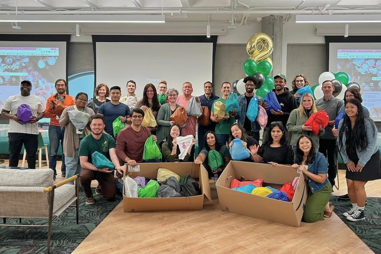 Group of volunteers in an office hold colorful drawstring bags, posing beside boxes of packed kits and green-and-gold balloons.