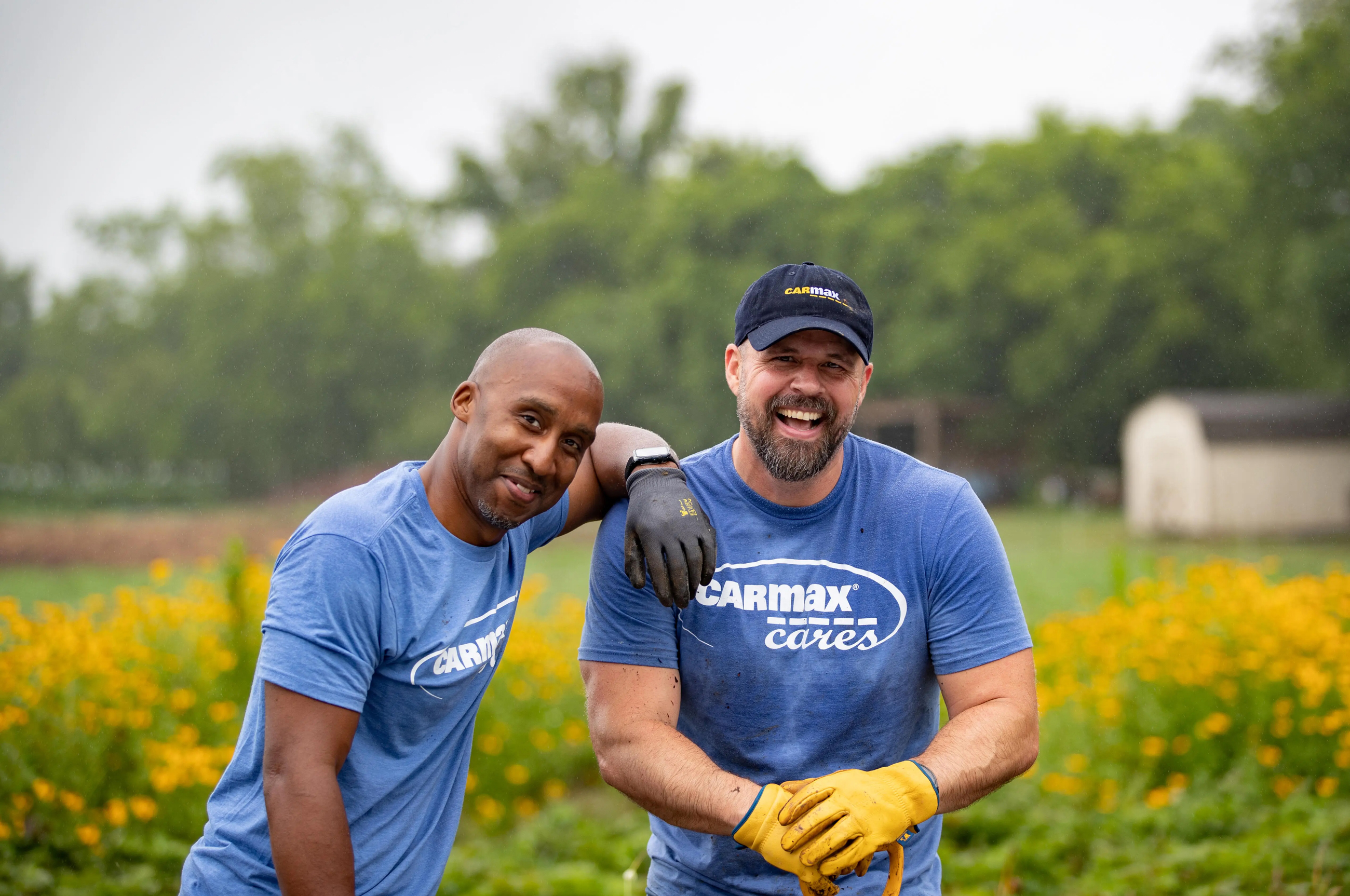 Two CarMax Cares volunteers in blue shirts smile in a flowered field; one in a cap with yellow gloves leans on a shovel.