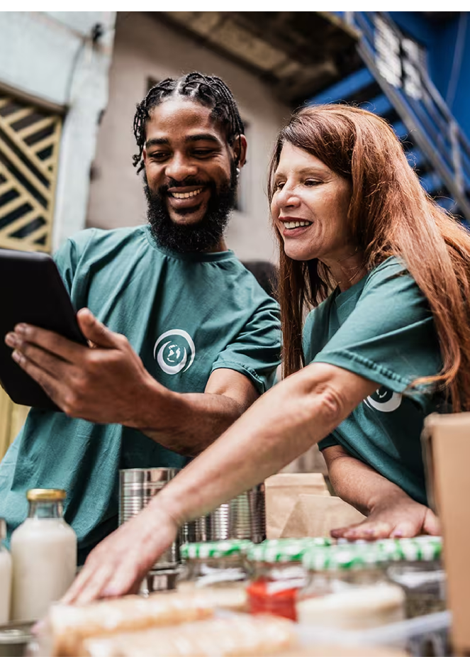 Two volunteers in green shirts smiling while checking a tablet at a food donation event.