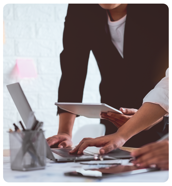 Two people stand at a desk with a laptop, tablet, and papers. One person holds a tablet while the others point at documents, suggesting a collaborative work or discussion environment.