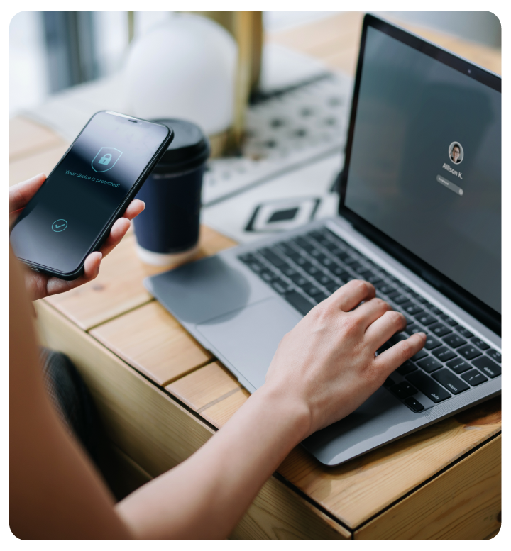 A person holding a smartphone showing a confirmation screen while logging in to a laptop, with a coffee cup and office desk in the background.