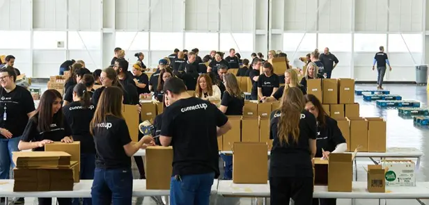 A large group of people from RTX in matching black shirts work together to pack cardboard boxes at long tables in a spacious, well-lit indoor area.