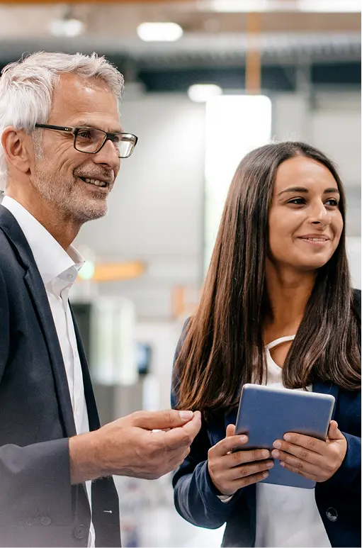 A man and a woman in business attire stand together indoors, smiling and engaged in conversation; the woman holds a tablet in her hands.