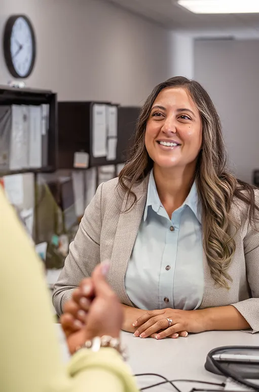Smiling woman in blazer talking to a person across the desk in a bank setting.