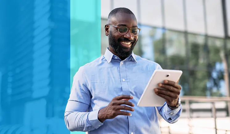 Smiling man in glasses and blue shirt using a tablet outdoors in front of a glass building.