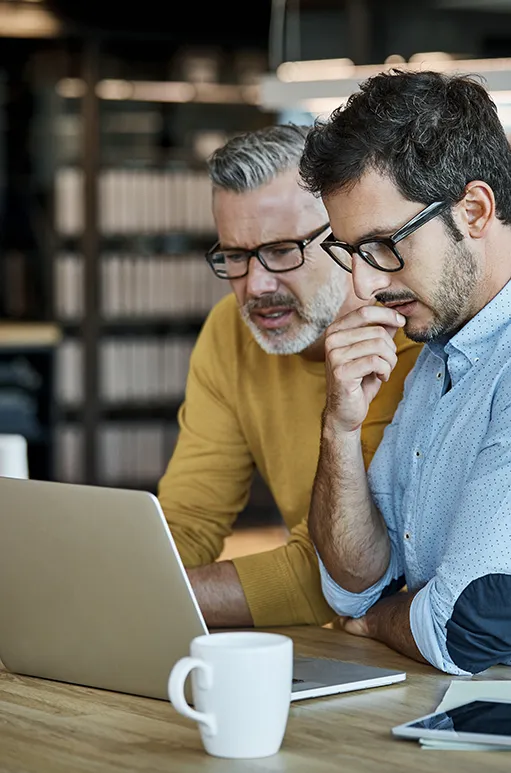 Two men wearing glasses attentively looking at a laptop screen in an office setting.
