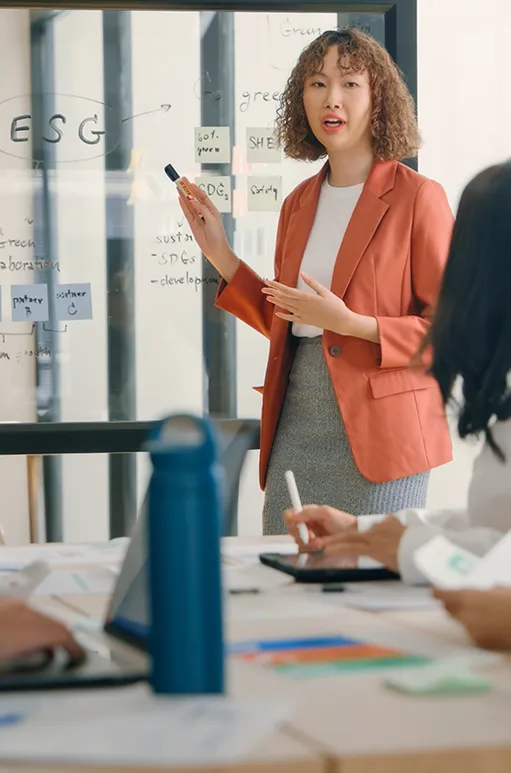 Woman in orange blazer explaining ESG concepts written on a glass board during a business meeting.