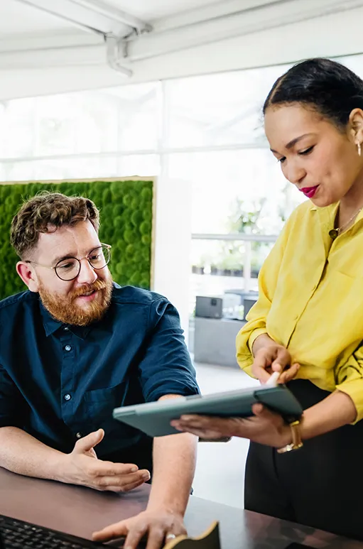 Man with glasses and beard sitting at a desk gesturing while a woman in a yellow blouse shows him something on a tablet.
