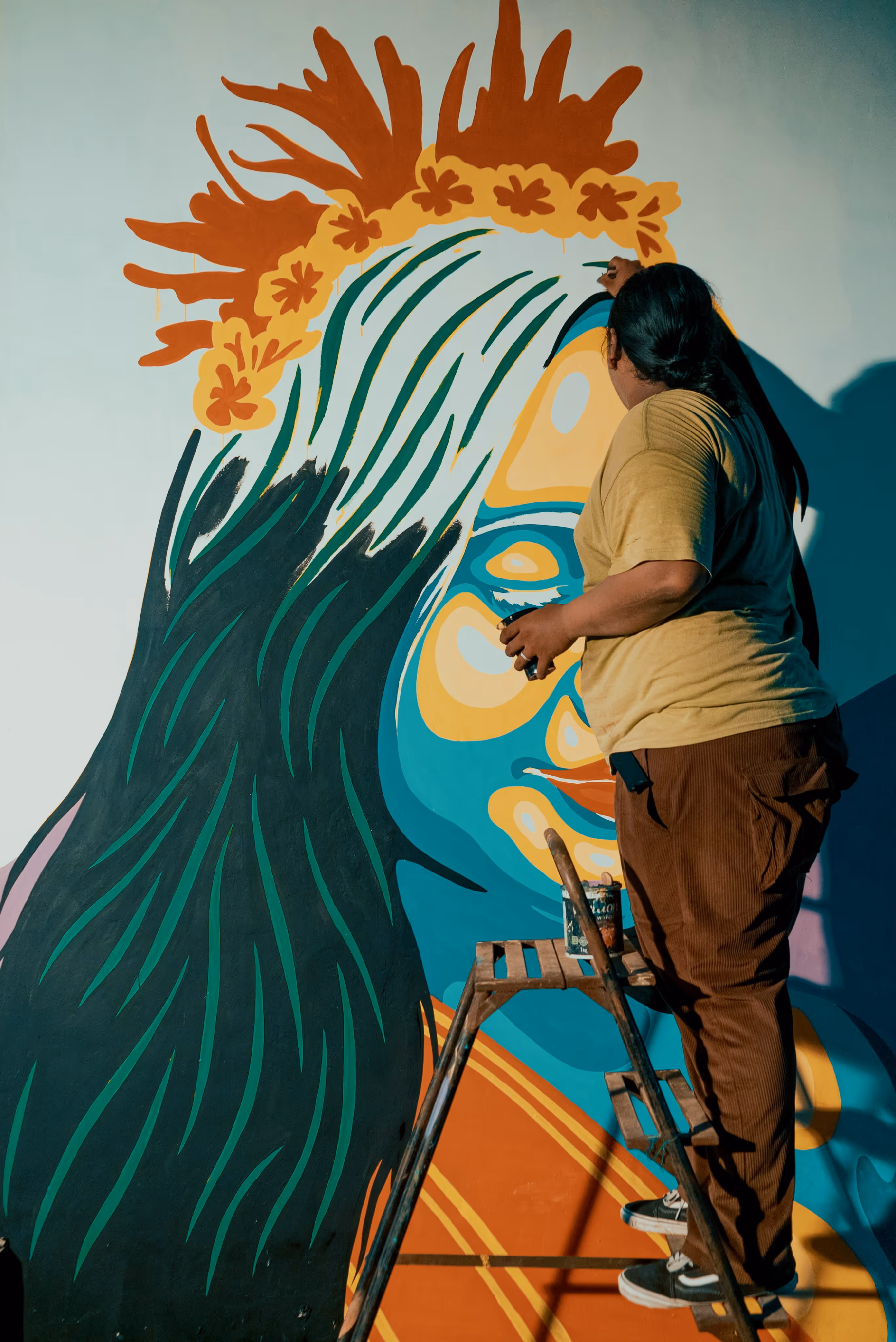 Person standing on a ladder painting a colorful mural of a stylized woman's face with flowers in her hair.