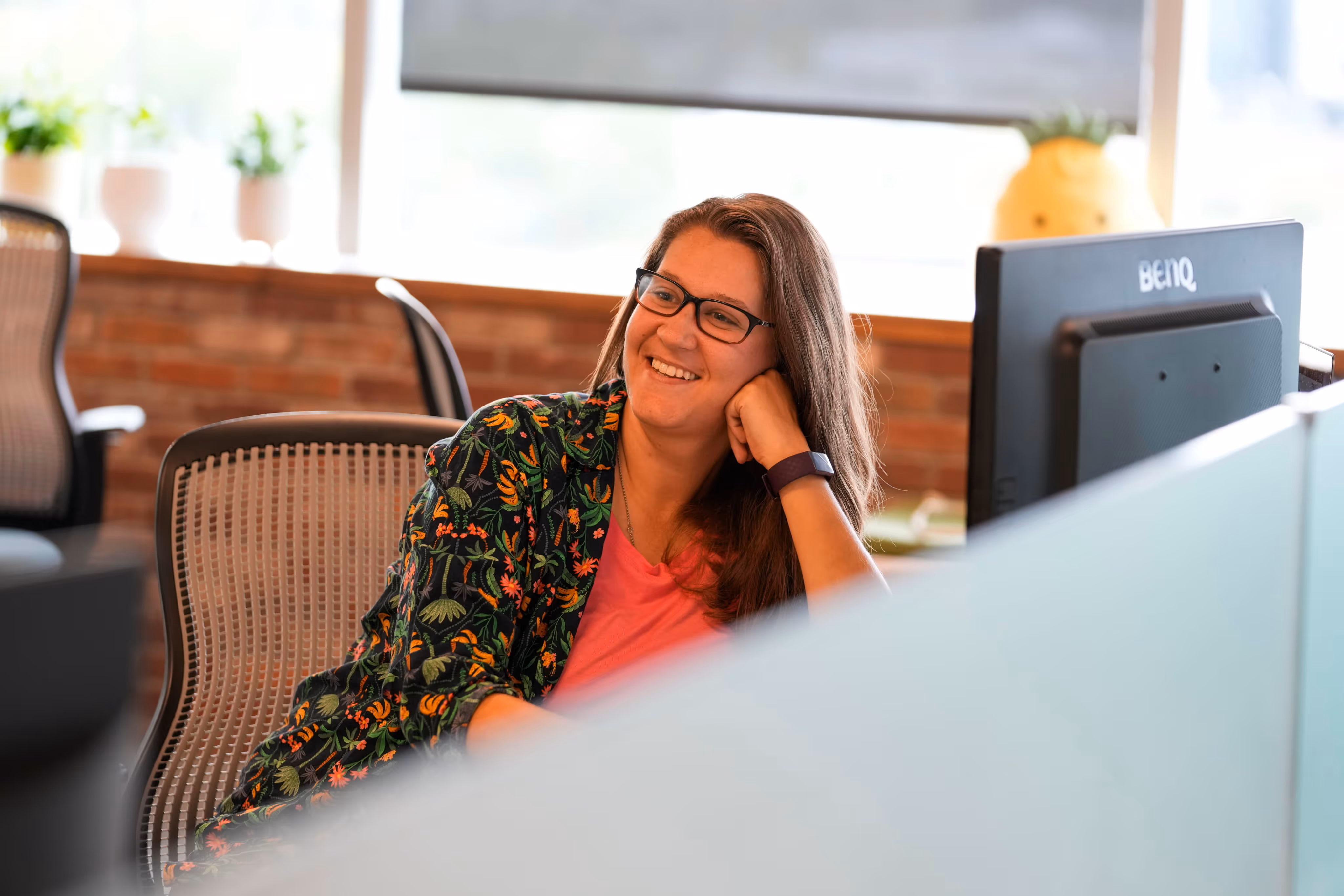 Smiling woman wearing glasses and floral top sitting in an office chair at her desk.