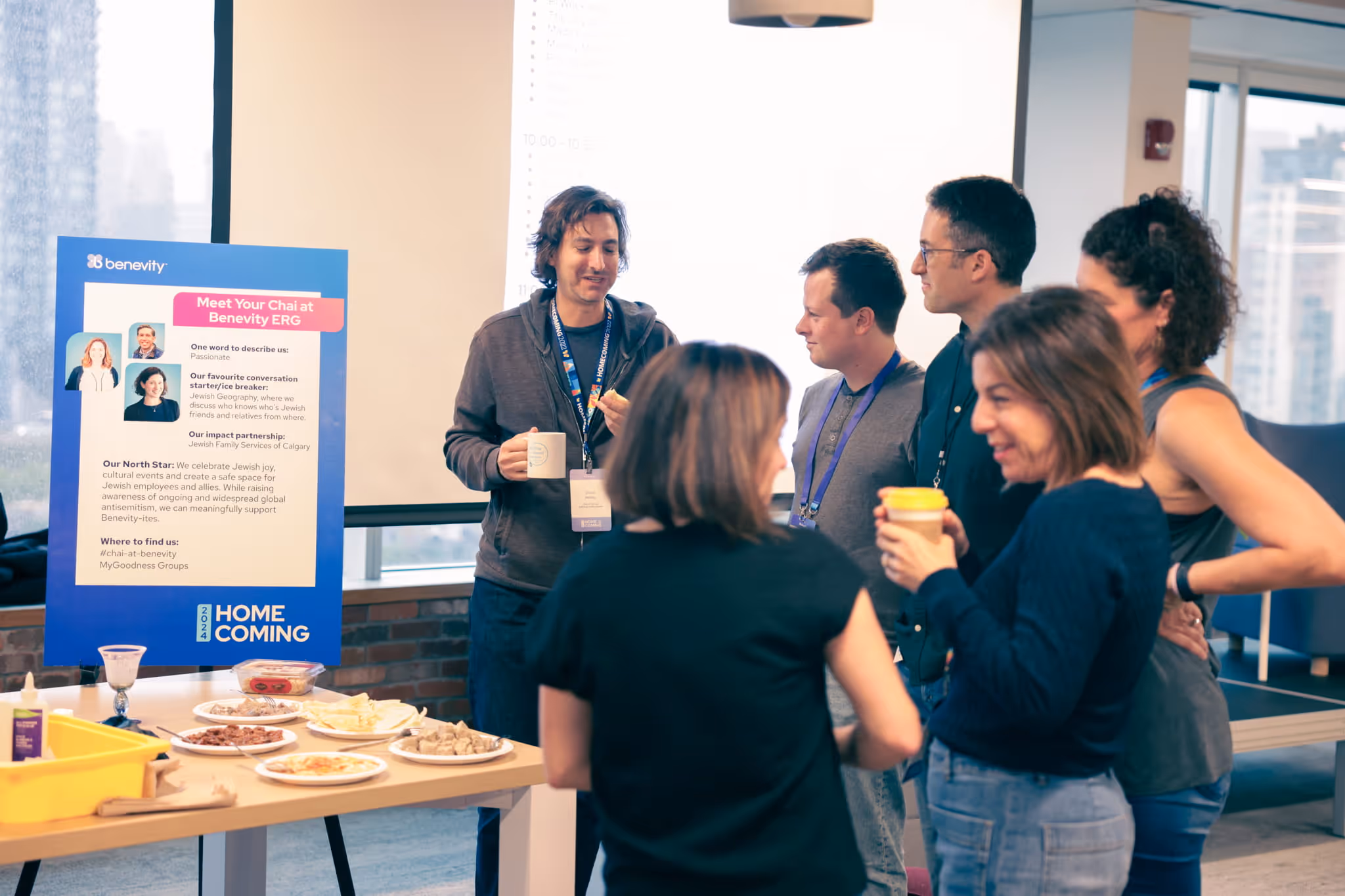 Group of six people socializing near a table with food and a Benevity ERG informational sign about Jewish cultural events at a 2024 Homecoming event.