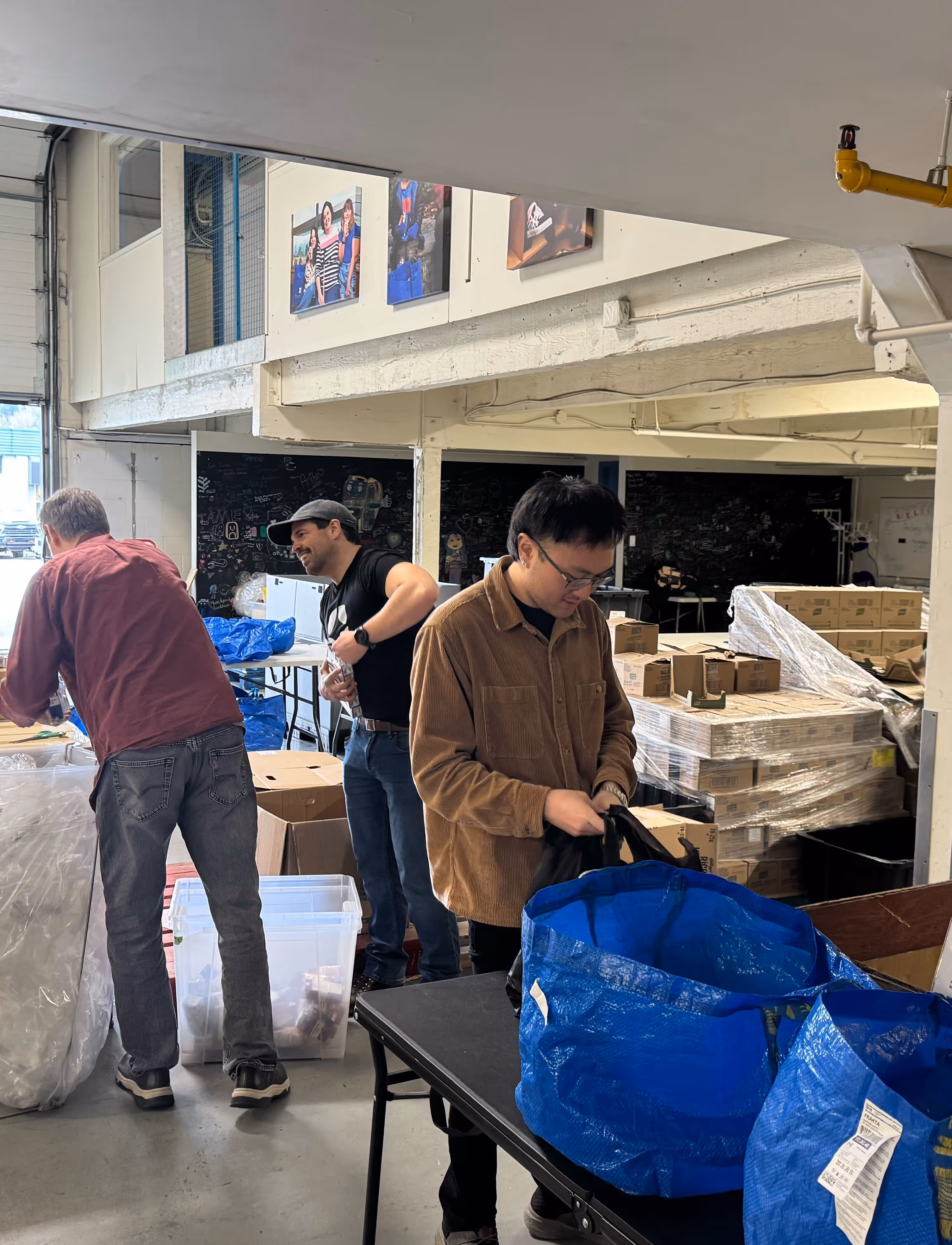 Three people packing items into blue reusable bags in a warehouse with stacked boxes and plastic bins around.