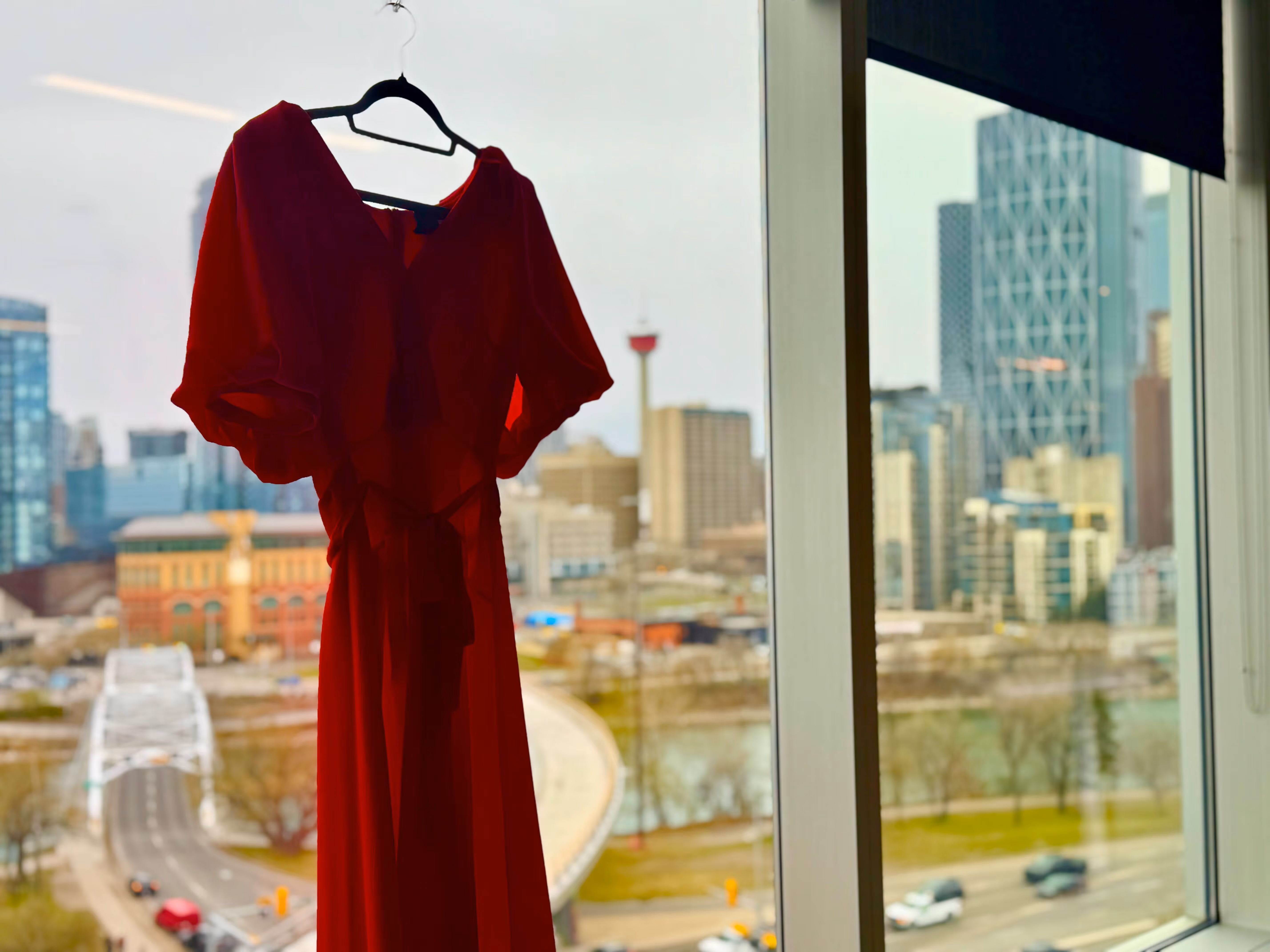 Red dress hanging on a black hanger in front of a large window overlooking a city skyline with buildings, a tower, a bridge, and a river.