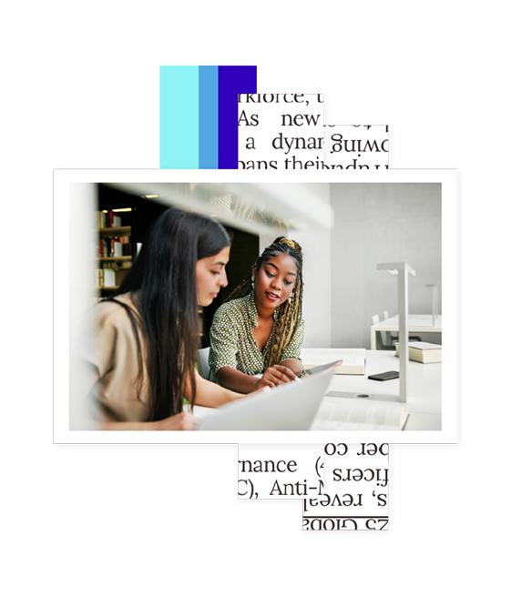 Two women working together at a table with a laptop and open books in a modern library or study area.