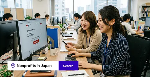 Two women smiling and working together at a computer in a busy office environment.