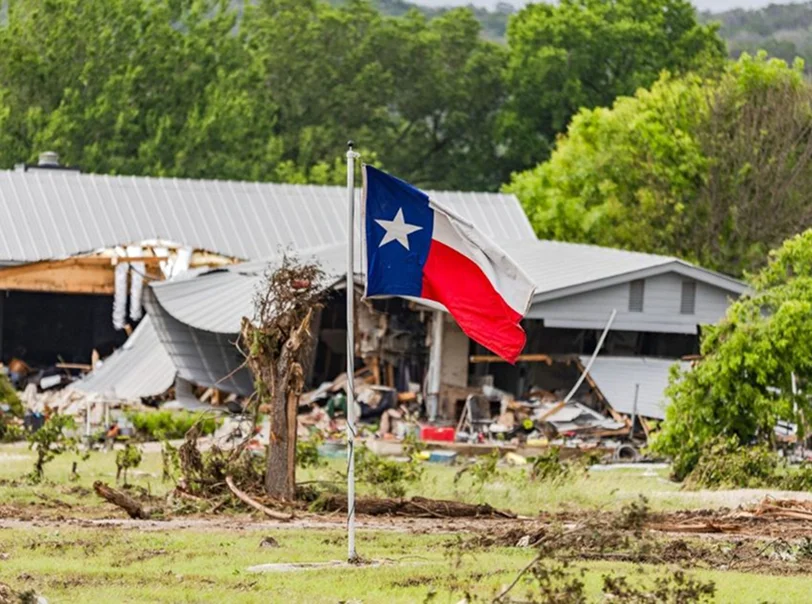 Texas flag on a pole in front of a damaged building with debris and broken trees.