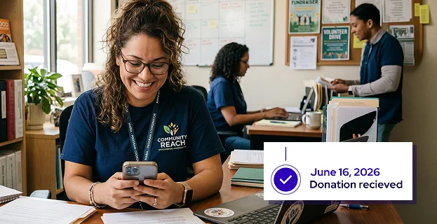 Smiling woman wearing a Community Reach shirt, looking at her smartphone, with two colleagues working in the background.