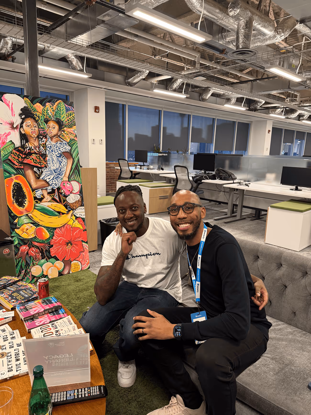 Two smiling men sitting closely on a gray couch in a modern office with colorful artwork and books on a nearby table.