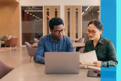 Two colleagues with glasses reviewing a document and laptop in a modern office meeting space.