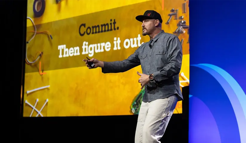 Man wearing a black cap and gray shirt speaking on stage with slide saying 'Commit. Then figure it out.'