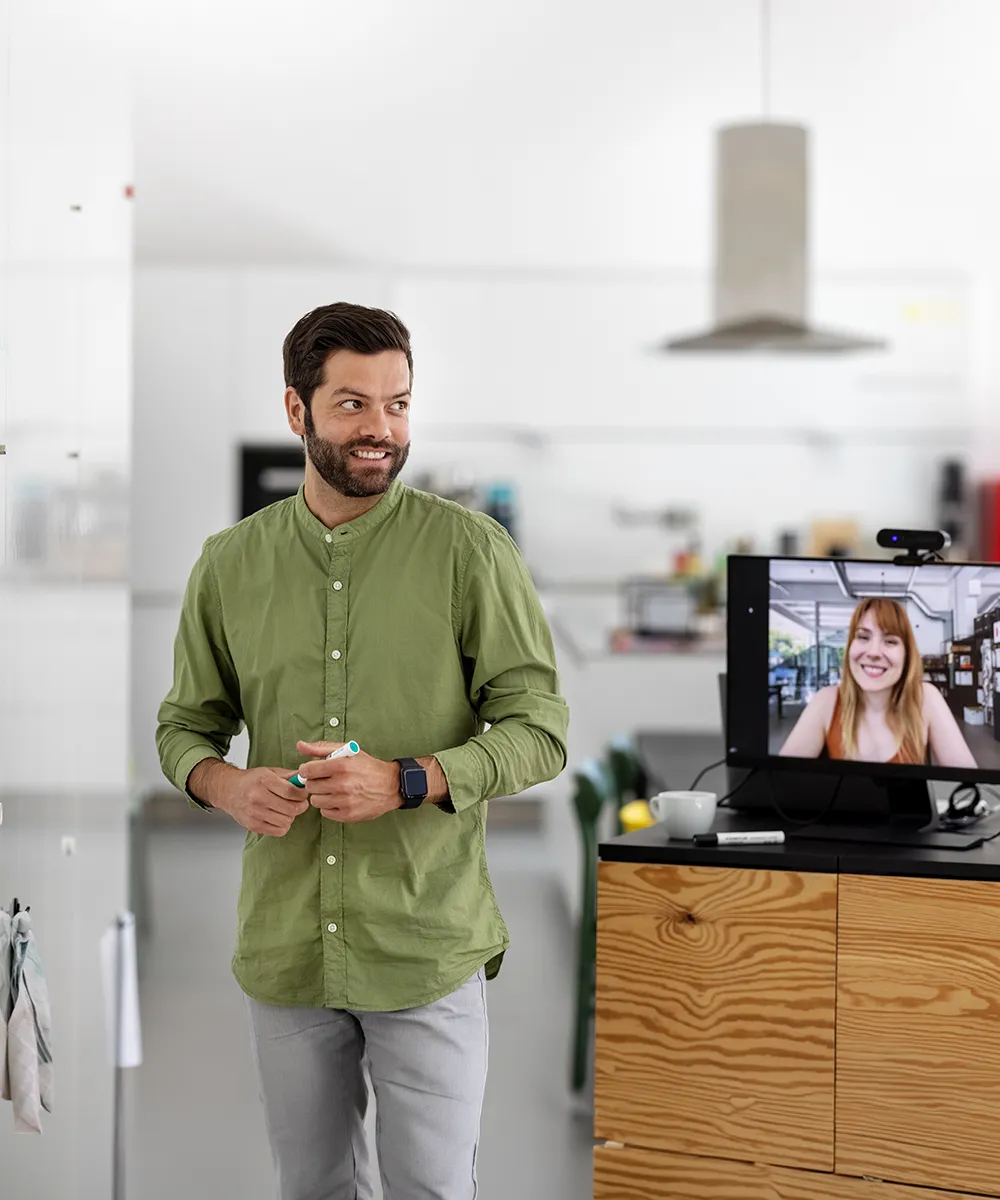 Man with beard and green shirt standing and holding a marker, looking to the side in a modern kitchen, while a woman with red hair smiles on a video call displayed on a monitor on a wooden counter nearby.
