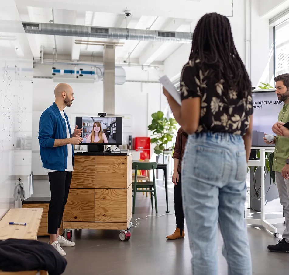 A diverse office team conducts a hybrid meeting with one man standing and talking near a monitor showing a woman on video call, while others stand nearby with notes and coffee in a bright modern workspace.
