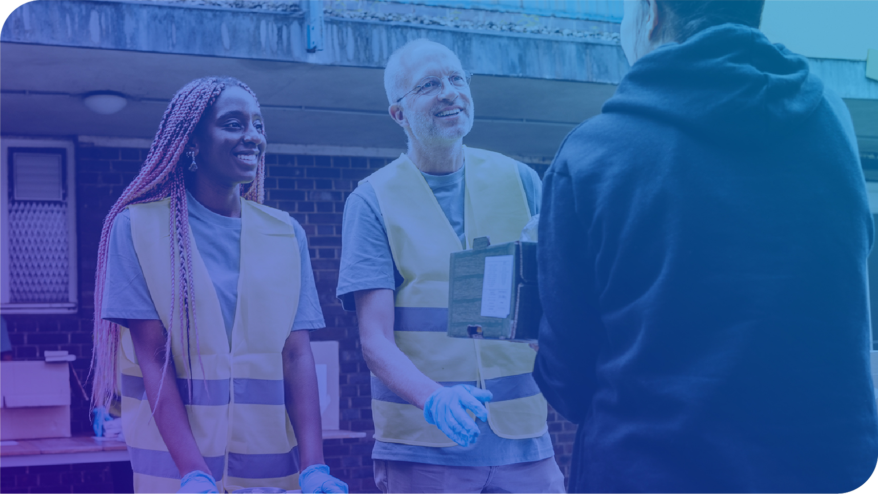 Two volunteers wearing yellow safety vests and gloves smile as they speak with a person holding a clipboard outdoors near a building.