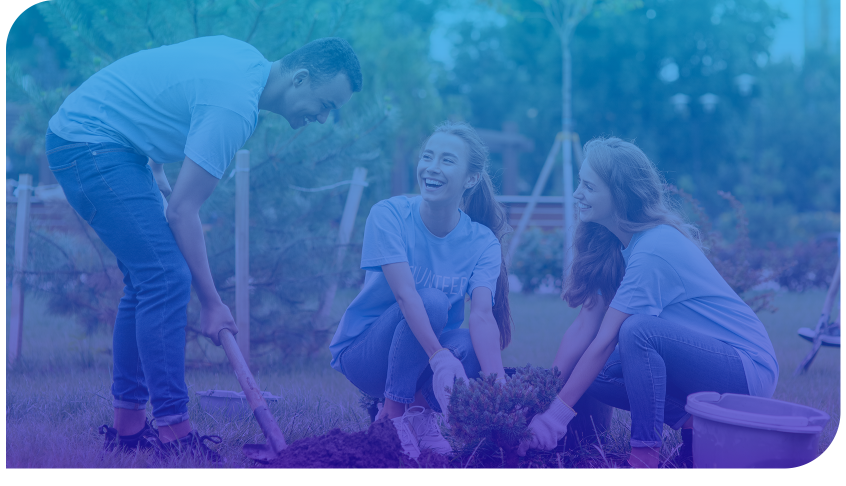 Three young people wearing blue volunteer shirts are outdoors, planting a small tree together and smiling. They appear happy and engaged in the gardening activity.