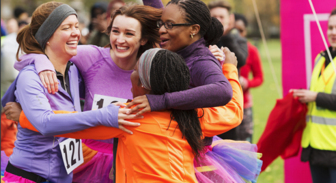 Four women wearing running gear and race numbers hug and celebrate at an outdoor event. They are smiling and appear joyful, with one woman wearing a rainbow tutu. A person in a yellow vest stands in the background.