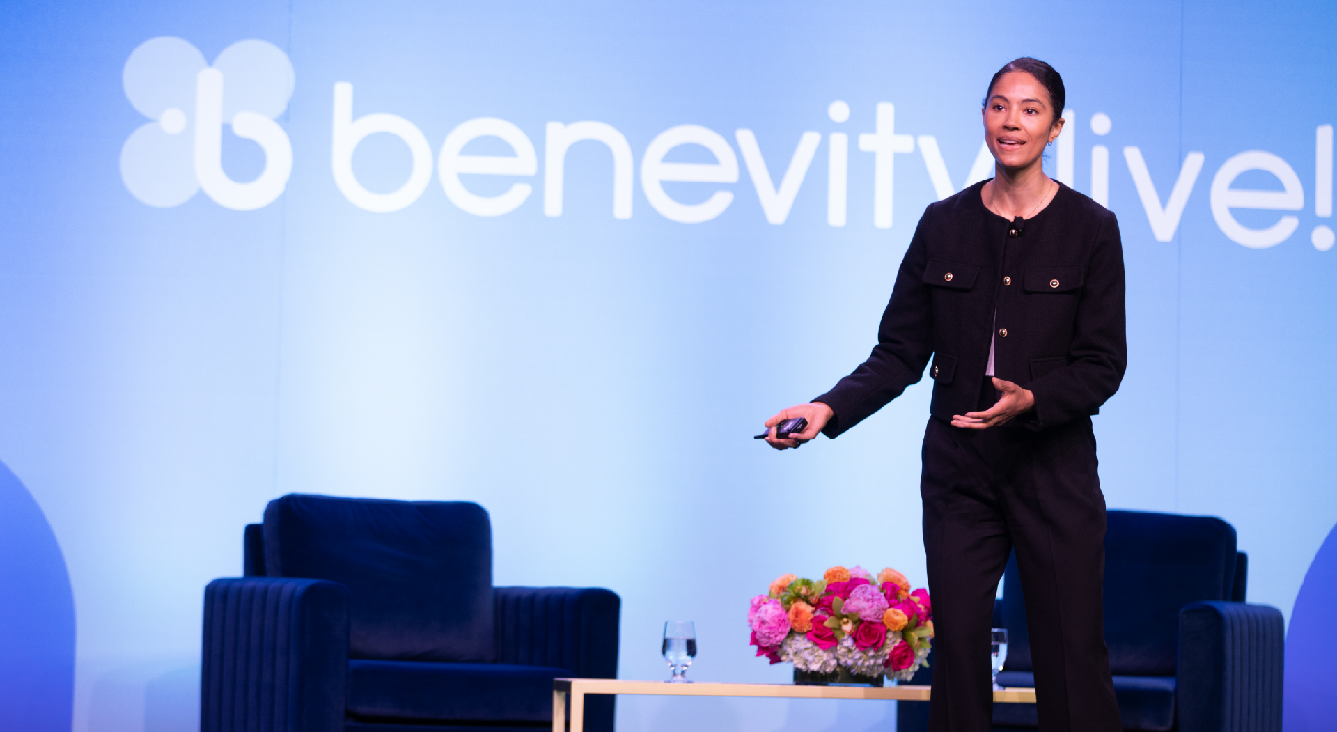 A woman stands on stage speaking at a Benevity Live! event, holding a presentation remote. Behind her are two blue chairs, a table with flowers and water, and a blue backdrop with the event logo.