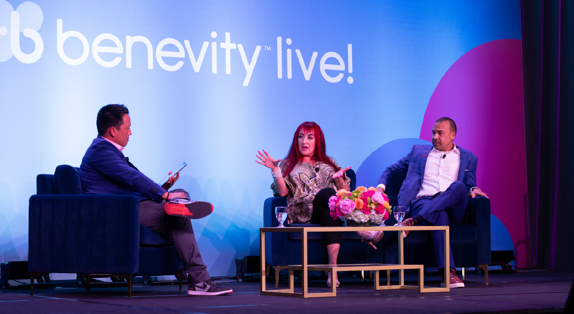 Three people sit on stage having a panel discussion at Benevity Live! conference. One person is speaking with expressive hand gestures, while the other two listen. A bouquet of flowers sits on the coffee table between them.
