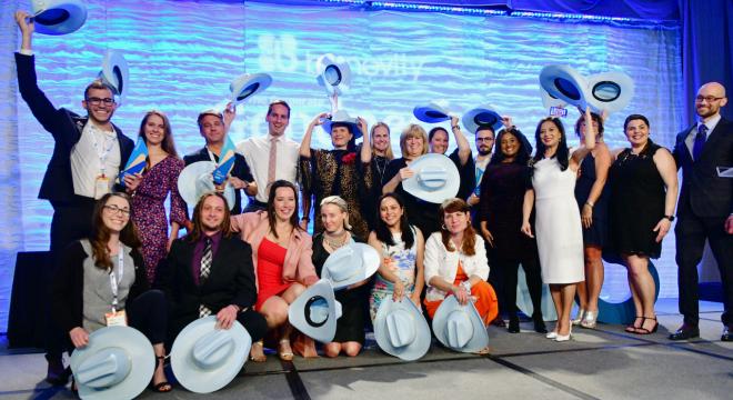A group of people dressed in formal attire pose together on stage, smiling and holding up light blue hats in celebration. The background features a blue-lit wall and a partially visible logo.