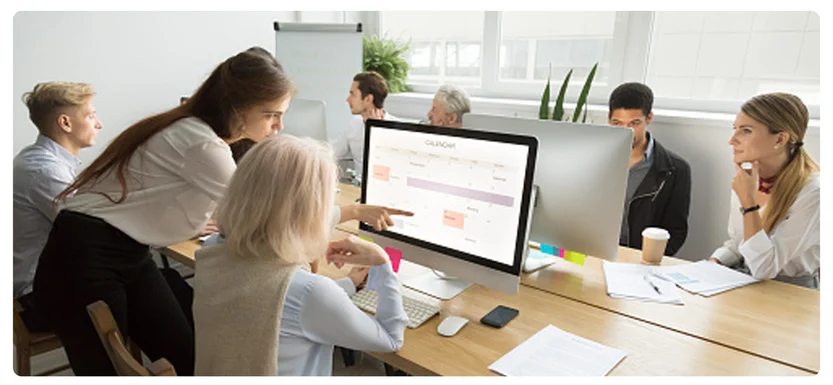 A group of people sit and stand around a long table in a modern office, collaborating and looking at a large computer screen displaying a chart or schedule.