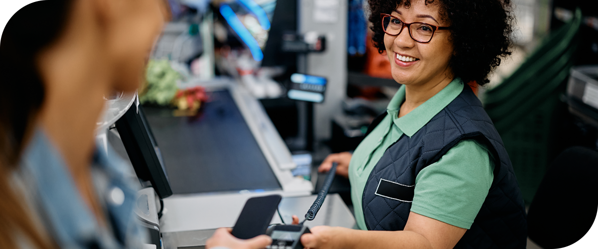 A smiling cashier in a green shirt and vest helps a customer pay with a smartphone at a checkout counter in a grocery store. Groceries and a payment terminal are visible.