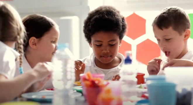 Three young children sit together at a table, eating and talking. They are surrounded by colorful cups and plates, and appear to be enjoying their meal in a bright, cheerful setting.